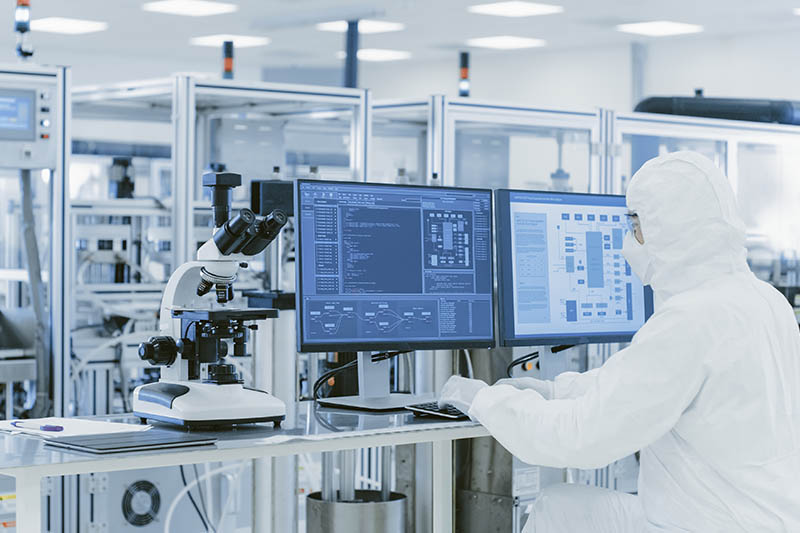 Pharmaceutical Manufacturing Facility In Laboratory Over the Shoulder View of Scientist in Protective Clothes Doing Research on a Personal Computer. Modern Manufactory Producing Semiconductors and Pharmaceutical Items.