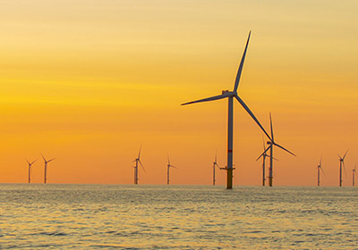 Energy Industry - Wind Turbine Offshore wind turbines at sunset, silhouetted against an orange sky and calm sea, representing environmental challenges like salt spray and corrosion.