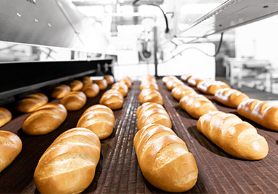 Food Industry - Bakery Freshly baked loaves of bread on a conveyor belt in a commercial bakery, illustrating a hygienic food processing environment with stainless steel equipment.